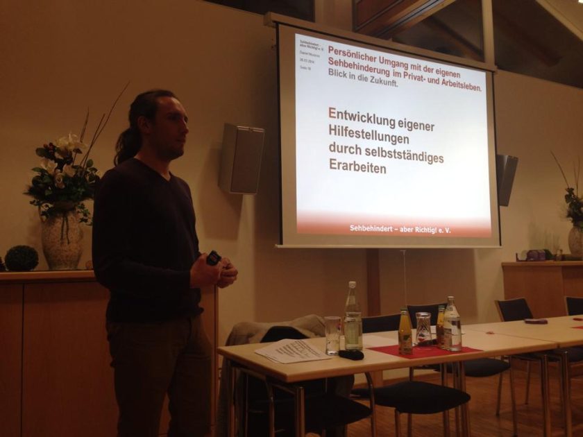 Eine Person hält einen Vortrag und im Hintergrund ist an einer Leinwand eine PowerPoint Präsentation zum Thema "Entwicklung eigener Hilfestellungen durch selbständiges Erarbeiten" zu sehen.